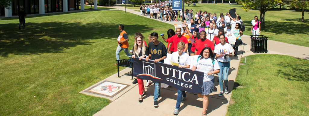 A group of students walking on a college campus on a sunny day. Several in front are holding a banner that reads "Utica College."